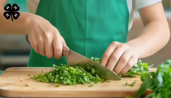 A child wearing a green apron using a knife on a cutting board to chop up food. A black 4-H clover in the upper right.