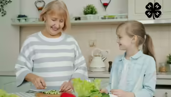 A grandmother and granddaughter in a kitchen. The grandmother chops vegetables on a cutting board. The black 4-H logo is in the upper right corner.