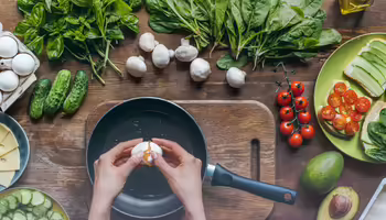 A person cracking an egg into a frying pan, surrounded by various ingredients to make a sandwich.
