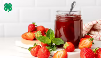 An open jar of strawberry jam with a spoon inside, surrounded by fresh whole and halved strawberries. A green 4-H logo in the top left corner.