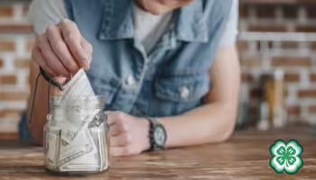 A person is placing folded dollar bills into a clear glass jar. A green 4-H clover in bottom right corner.