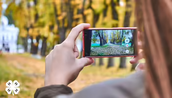A person using a cell phone to take an image of a fall scenery. A white 4-H clover is on the bottom left.