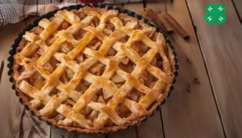 A baked apple pie with a golden-brown lattice crust in a metal pan on a wooden table. The green 4-H logo is in the right upper corner.