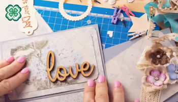 A person arranges a scrapbook page surrounded by craft supplies like lace, paper flowers, and ribbons. A green 4-H clover in the upper left corner.