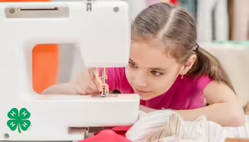 A girl focusing on the needle of a sewing machine. The green 4-H clover is in the bottom left corner.