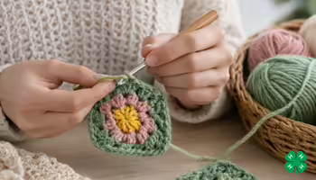 A person's hands using a crochet hook to craft a colorful floral square, with yarn balls in a woven basket. A green 4-H clover in bottom right corner.