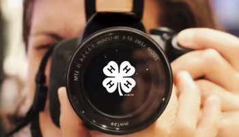 A person holding a camera up like they are taking a photo. A white 4-H clover in the middle of the camera lens.