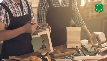 An adult and a child wearing aprons work together at a woodworking bench filled with tools. The 4-H logo is in the top right corner.