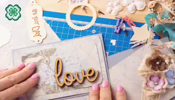 Scrapbooking craft table with cardstock, wooden embellishments, and the 4-H logo.