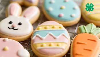 Iced sugar cookies in the shapes of a bunny, eggs, and a carrot on a wire cooling rack with a green 4-H logo in right upper corner.