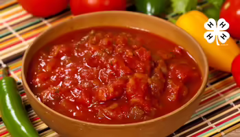 A bowl of salsa of sitting on a placemat with peppers and tomatoes around the bowl.