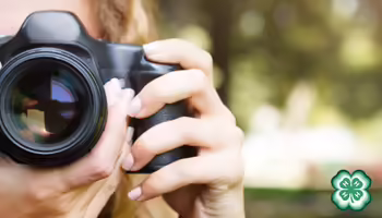 Close-up of a person holding a camera to their eye, with a green 4-H clover logo in the bottom right corner and a blurred outdoor background.