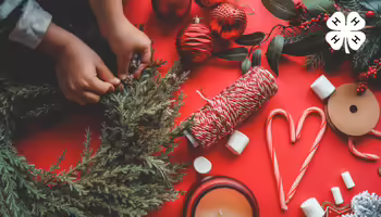 Hands assembling a holiday wreath alongside red ornaments, candy canes, and craft supplies on a red background with a 4-H logo.