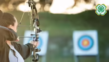 A person aims a bow at a target at s outdoor archery range. A 4-H logo in the top right corner.