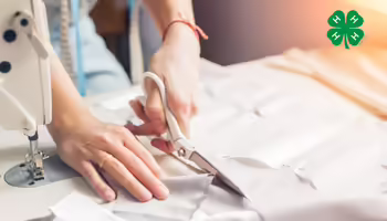 A person cutting fabric next to a sewing machine. A green 4-H clover in the upper right corner.