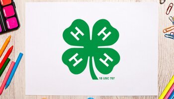 White paper in the middle with a green 4-H clover on it. Art supplies such as paint, crayons, colored pencils, pens, and paperclips surround the piece of paper.