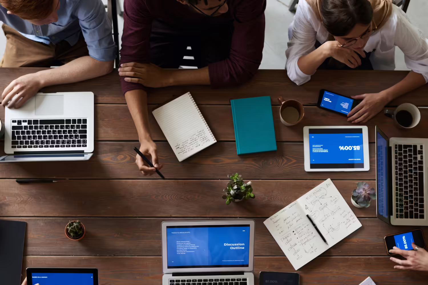 view from above of a meeting full of notebooks, computers, and cell phones and different arms reaching in
