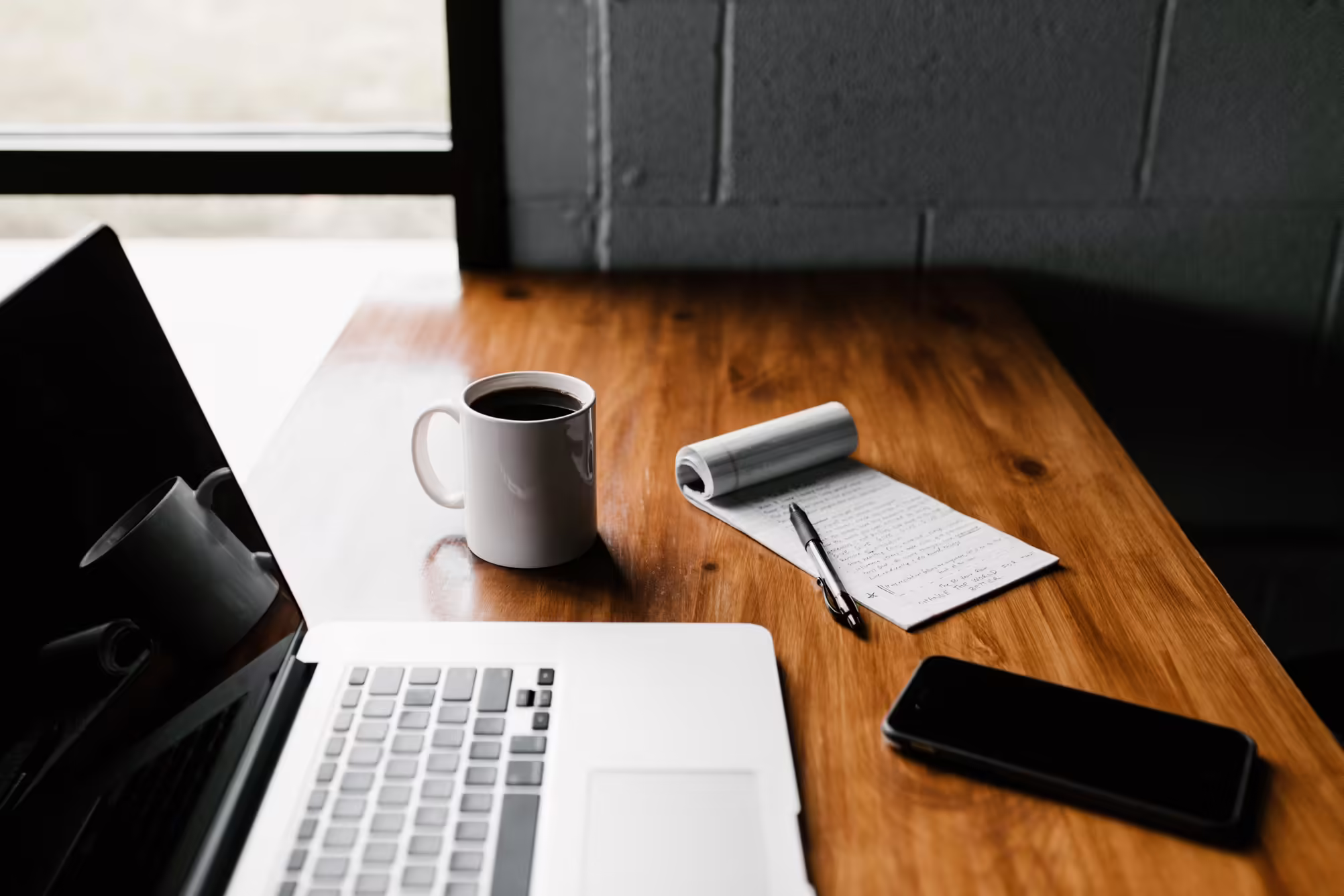 A laptop next to a cup of coffee, notepad with a pen and a cell phone.