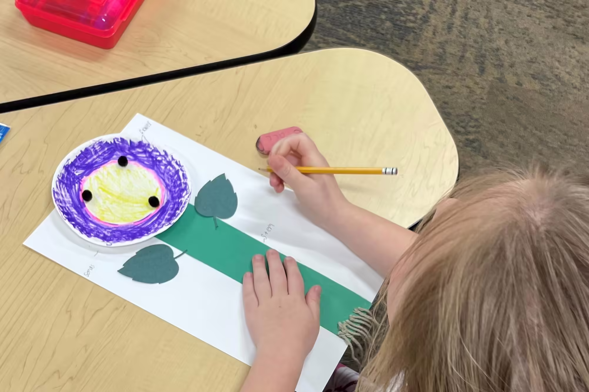 student sits at a table, creating a flower from paper and a colored plate