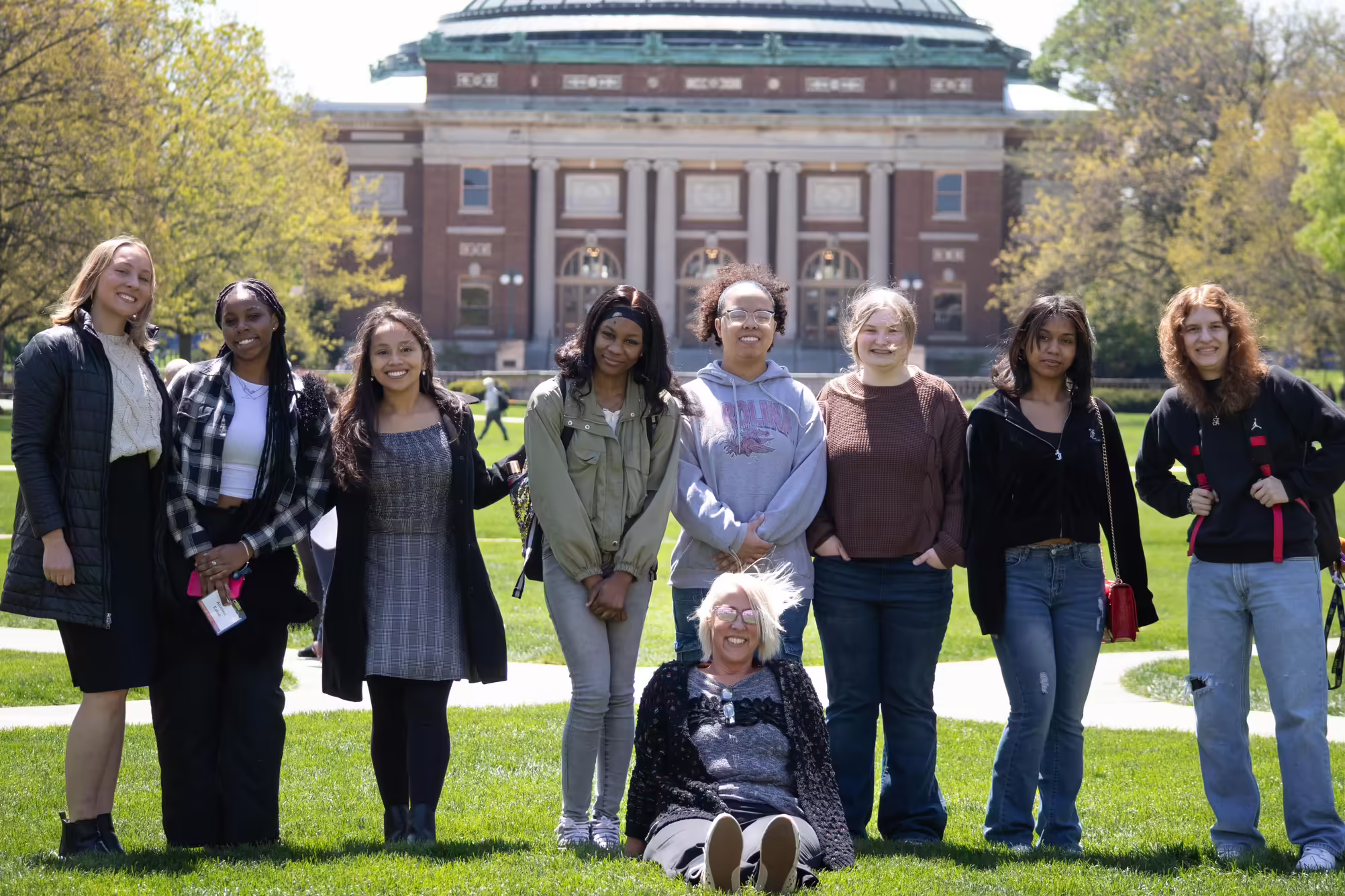 9 young adults sit and stand outside on the quad at the University of Illinois