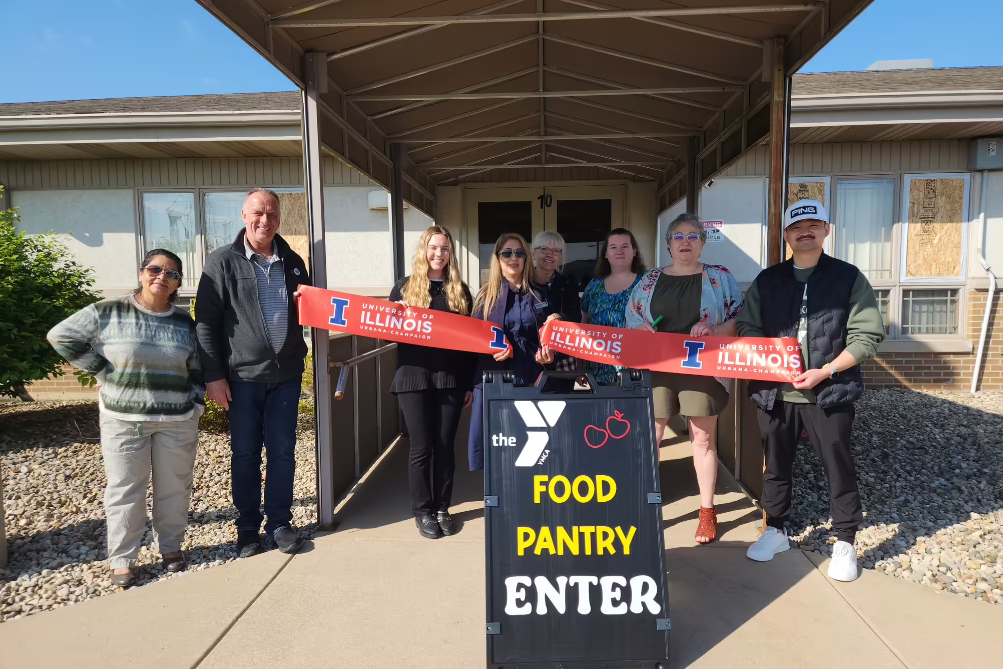 A group of community leaders during a food pantry grand opening pose with a cut ribbon. A sign in front of them reads "Food Pantry Enter"