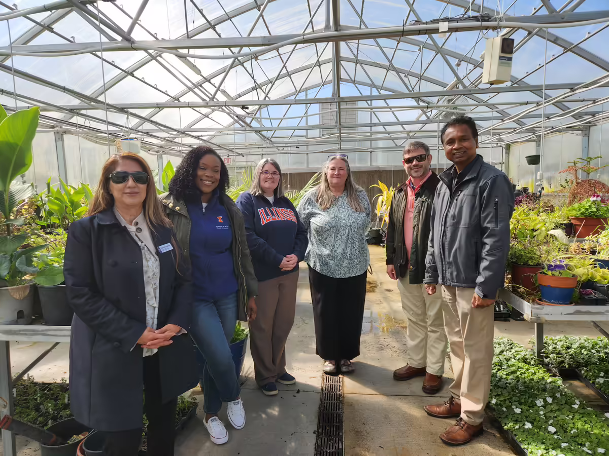 Six people, some wearing branded University of Illinois apparel, stand inside a greenhouse