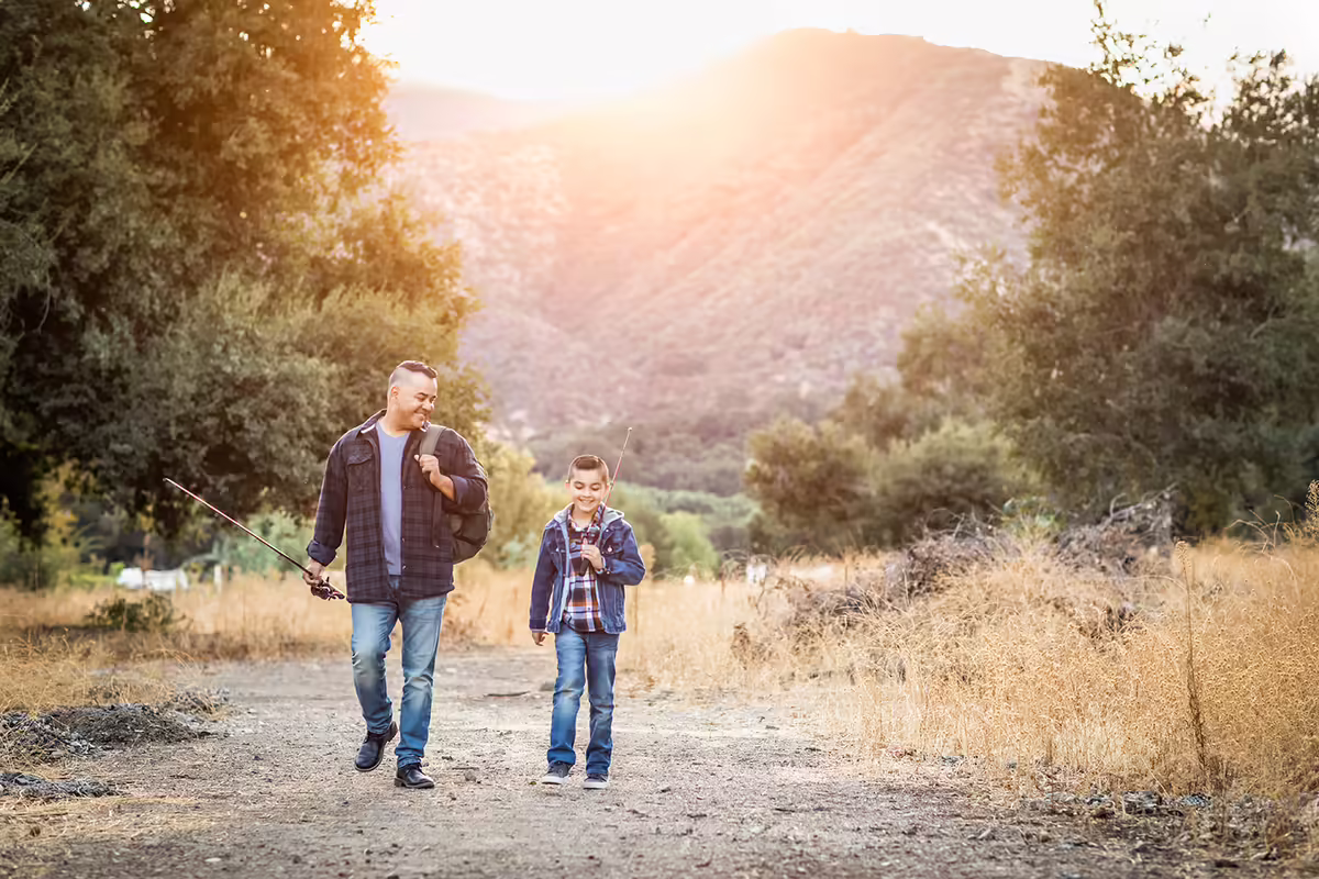a father and son walk down a path in a nature area with fishing poles