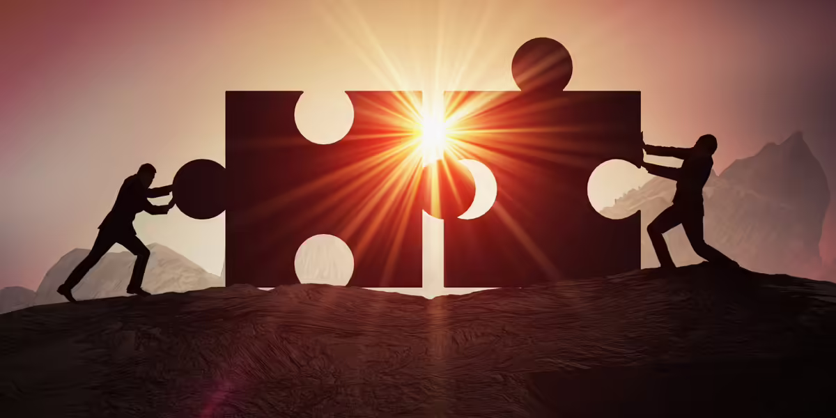Two silhouettes pushing two giant puzzle pieces together in front of a setting sun.