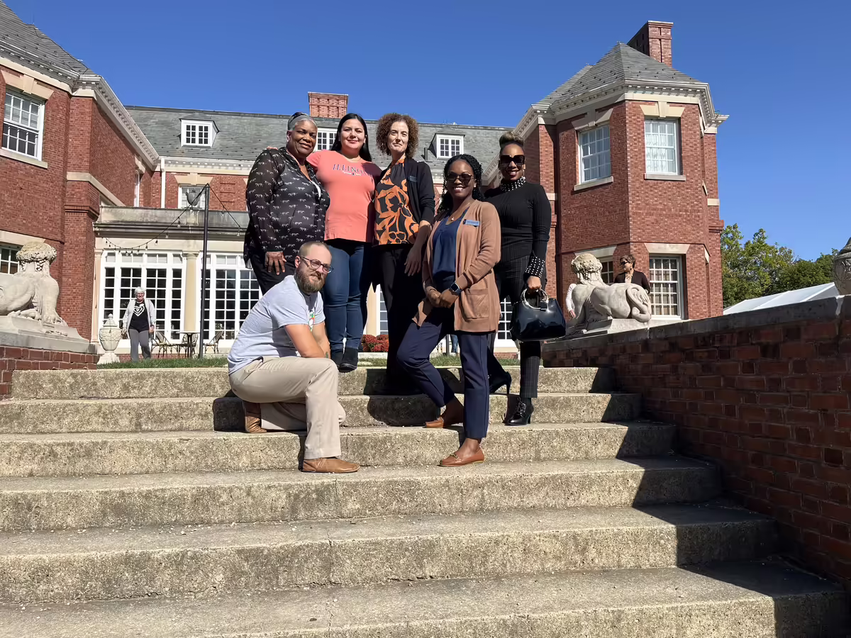 6 adults standing/kneeling on concrete steps, outside, in front of a brick building