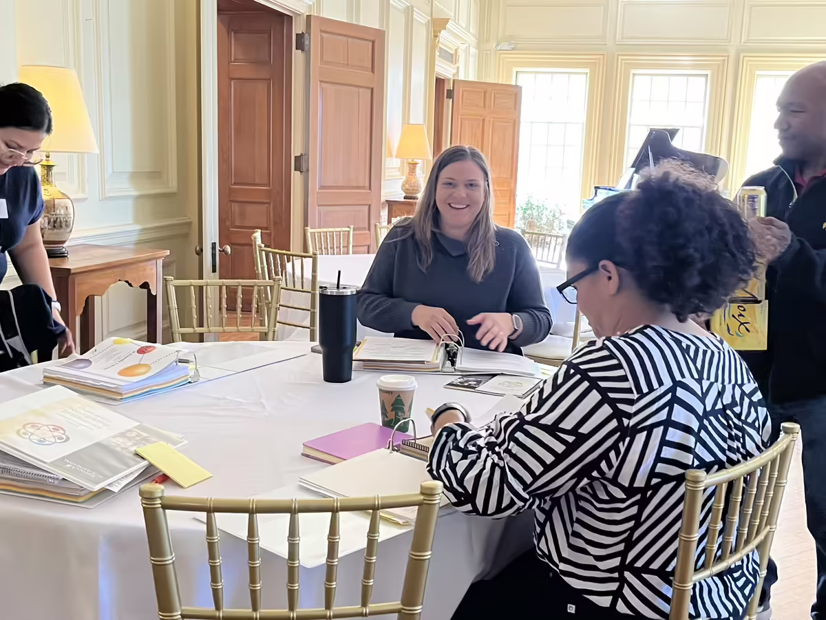 2 adult women sitting at a round table. One is looking at the table & materials, the other is looking at the camera smiling