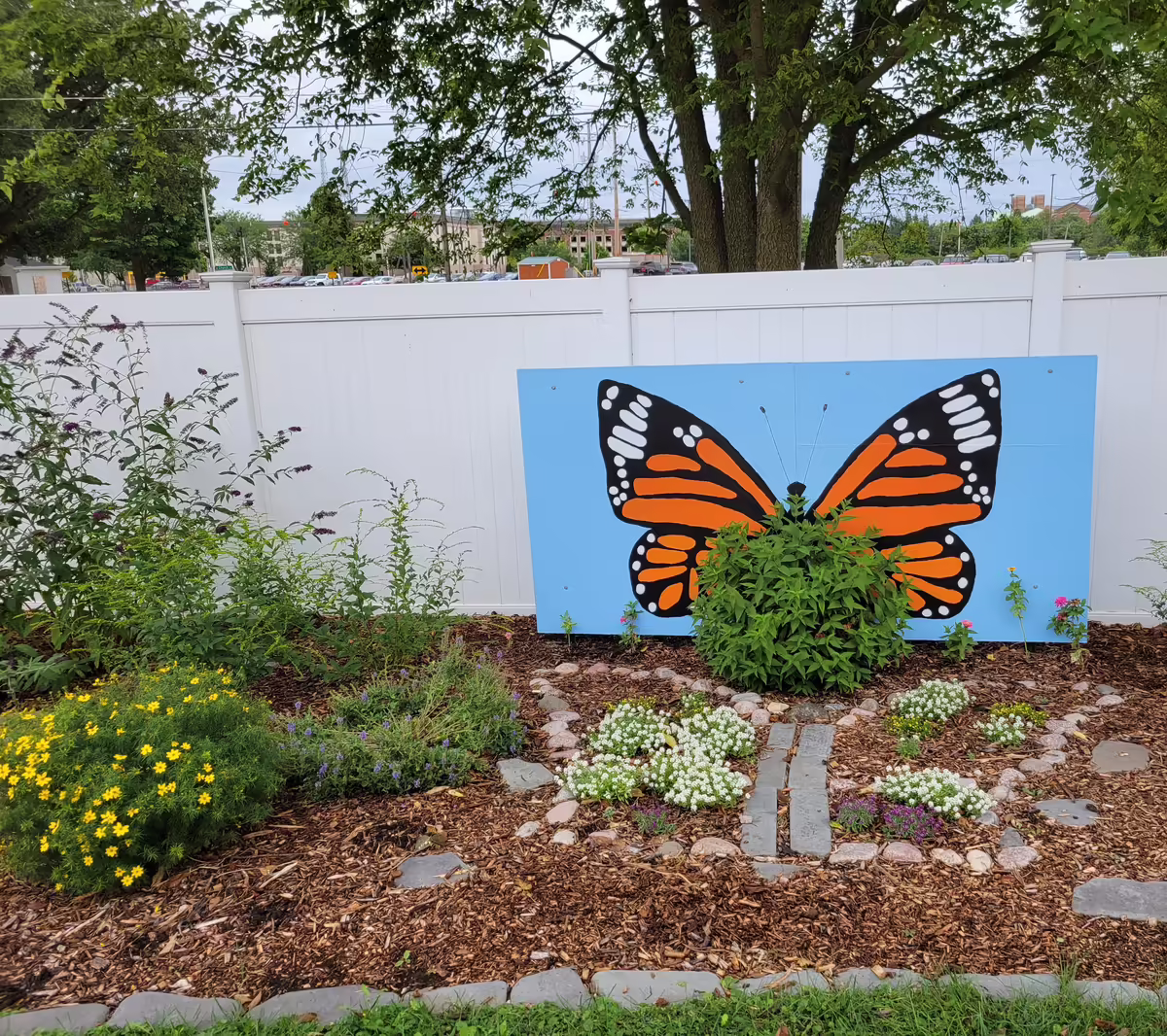 A garden of shrubs and decorative stones in a butterfly shape in front of a mural of a monarch butterfly.