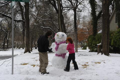 Man and female child build a snowman in residential neighborhood among trees