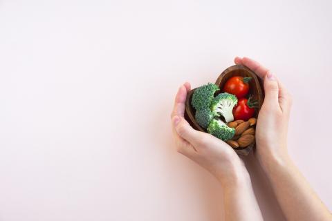 A hand holding a heart shaped wooden bowl with broccoli, tomatoes, and nuts in it.