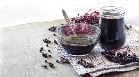 A bowl and jar of elderberry syrup, with elderberries surrounding them.