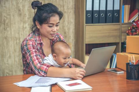 Young adult working at laptop holding child.