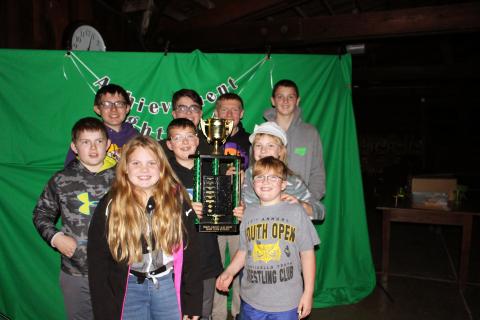 Nine kids standing in front of a green 4-H backdrop holding a trophy. 