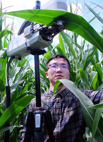An asian man stands in a corn field with a tool that is attached to a corn leave. 