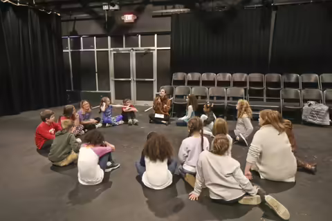 A group of kids and 1 adult sitting in a circle on a stage.