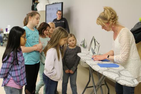 A woman working with fabric on an ironing board while 6 children observe.