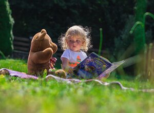 child sitting on grass holding a book with her toy teddy bear