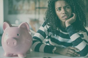 woman with questioning look on face sitting next to piggy bank