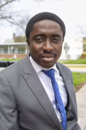 Nigerian man posing for an upper body headshot wearing a grey suit with a white shirt and blue tie.