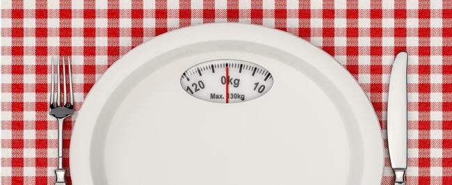 white plate with a scales on upper portion of plate, sitting on a red gingham background with a knife and fork