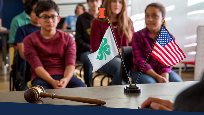 Children sitting in chairs while the 4-H flag, and American Flag, and a gavel are displayed on a table. 
