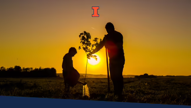 A young boy using a watering can to water a tree that an older man is holding.