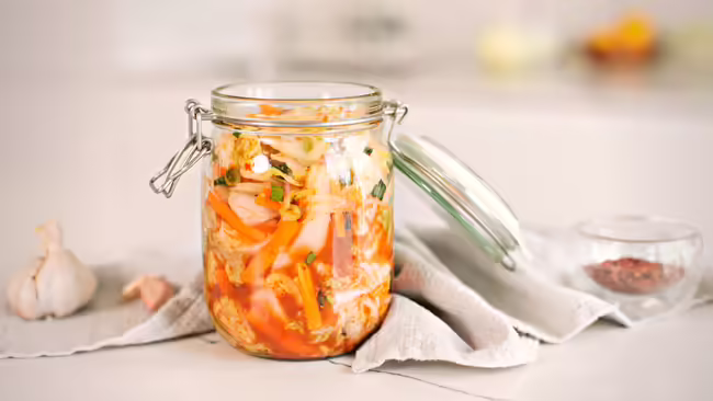 Image of a jar of kimchi on a cloth napkin next to a garlic clove and a small bowl of spices