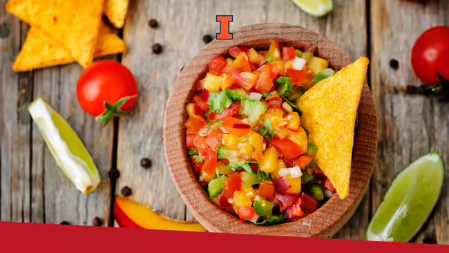 Apple mango salsa in a wooden bowl placed on a wood table with tomatoes, limes, and chips placed around the bowl on the table.