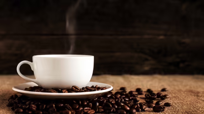 A cup with steam coming out of it sitting in coffee beans on a wooden table.