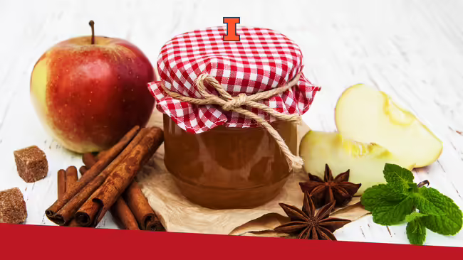 Apple jam in a jar and an apple, apple slices, cinnamon sticks, sugar and spices sitting on a wooden table.