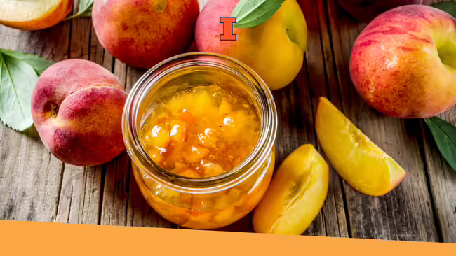 Peach jam in a jar sitting on a wooden table with whole peaches and half peaches surrounding the jar